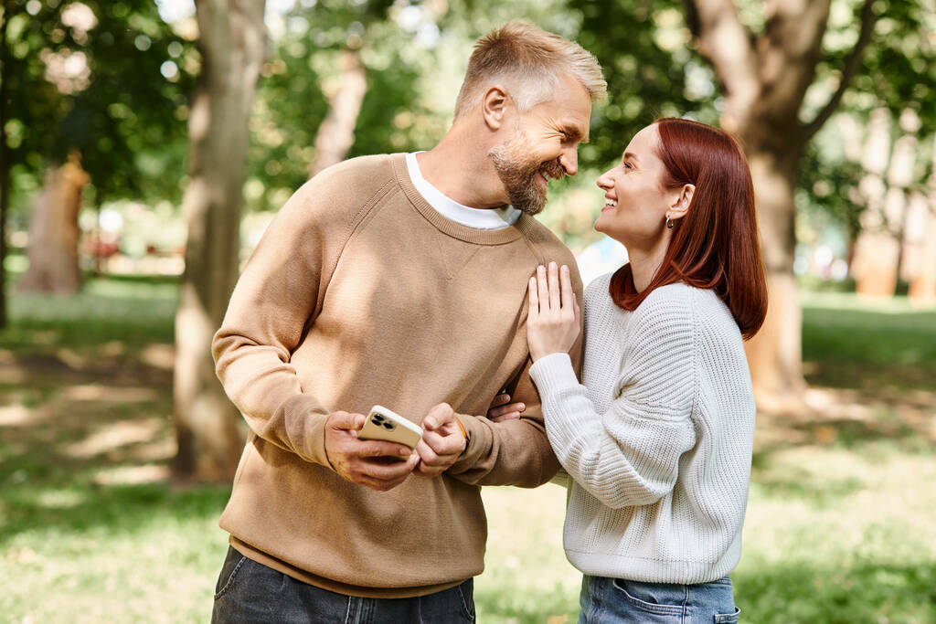 Um homem e uma mulher de traje casual abraçam enquanto estão juntos em um parque vibrante. - Foto, Imagem