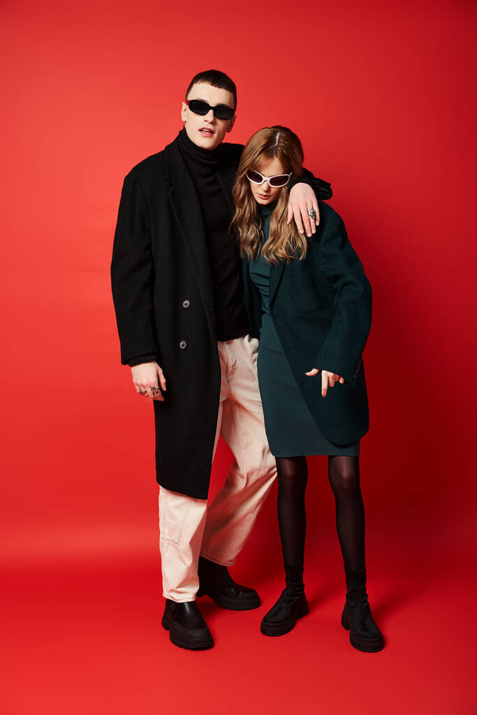 модна красива пара в стильних пальто з сонцезахисними окулярами позує разом на червоному тлі - Фото, зображення
