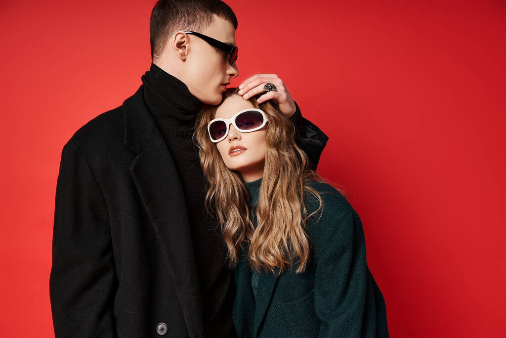 fashionable young couple in stylish coats with trendy sunglasses posing together on red backdrop - Photo, Image