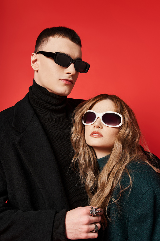 modna młoda para w stylowych płaszczach z modnymi okularami przeciwsłonecznymi pozującymi razem na czerwonym tle - Zdjęcie, obraz