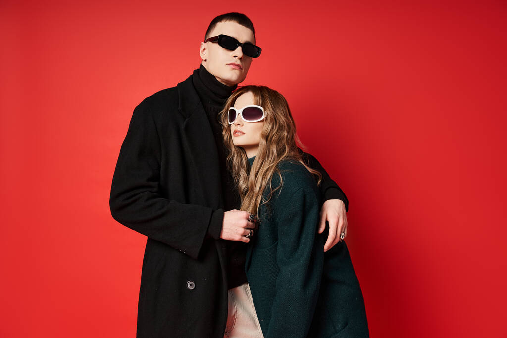 sophisticated young couple in stylish coats with sunglasses posing together on red backdrop - Photo, Image