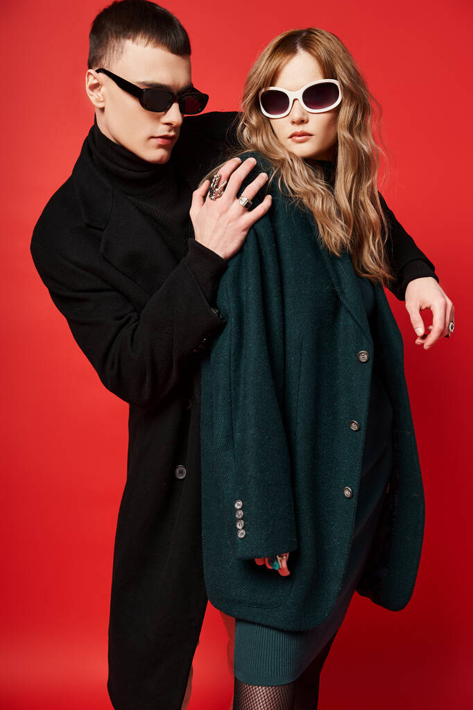 добре одягнена молода пара в стильні пальто з сонцезахисними окулярами позує разом на червоному тлі - Фото, зображення