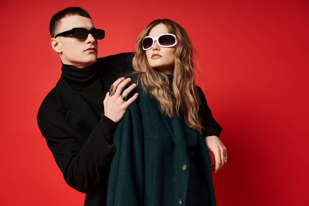 élégant jeune couple en manteaux élégants avec des lunettes de soleil à la mode posant ensemble sur fond rouge - Photo, image