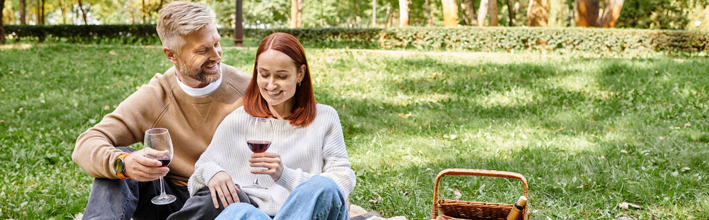 Volwassen man en vrouw zittend op een deken, wijnglazen vasthoudend in een park. - Foto, afbeelding