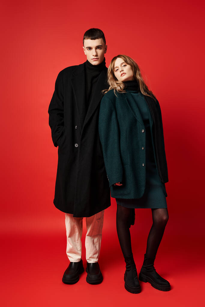 appealing loving couple in sophisticated coats looking at camera on red vibrant background - Photo, Image