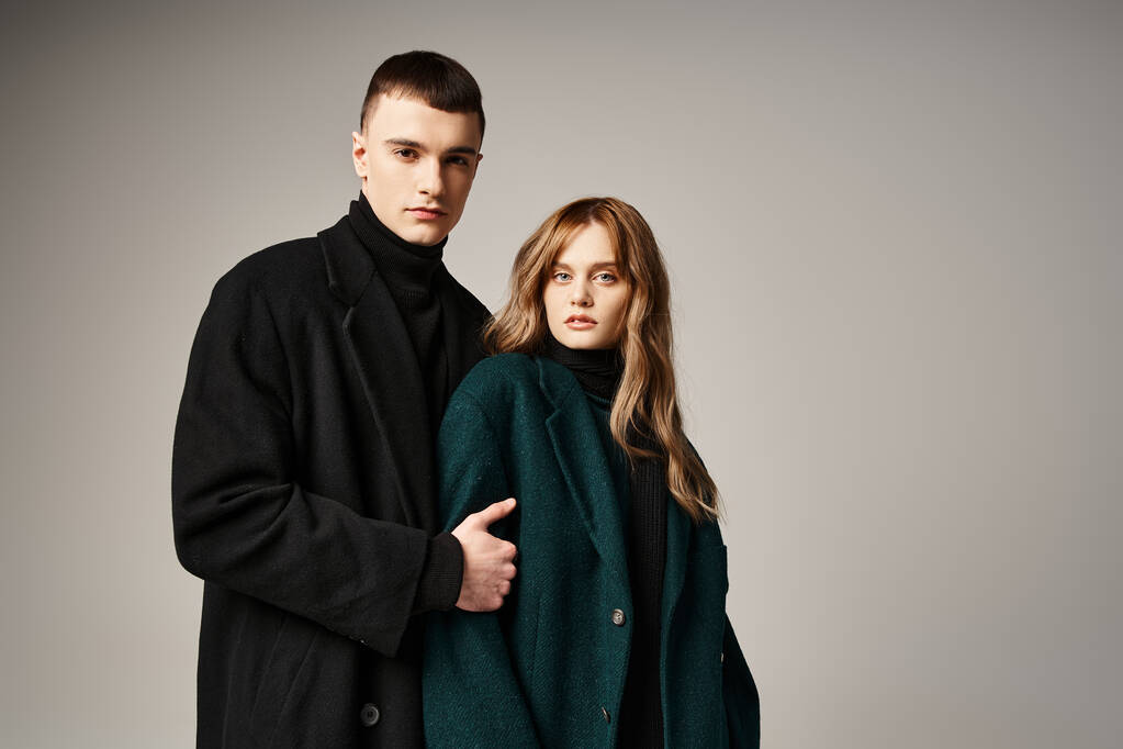 coppia alla moda in eleganti cappotti in posa insieme su sfondo grigio e guardando la fotocamera - Foto, immagini