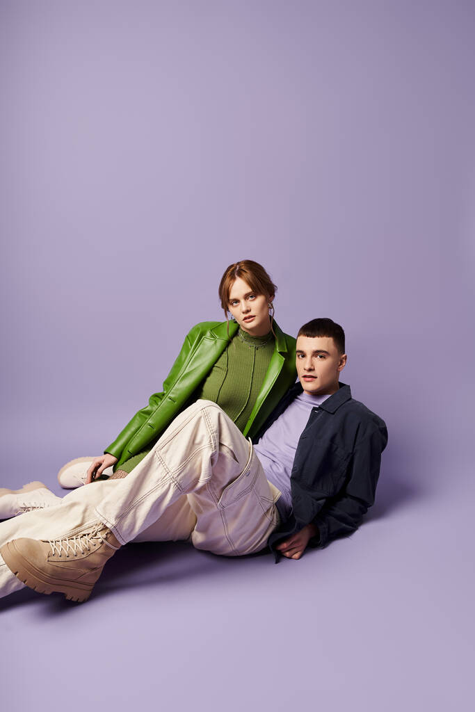 coppia ben vestita in abiti vibranti seduti sul pavimento e guardando la fotocamera sullo sfondo viola - Foto, immagini