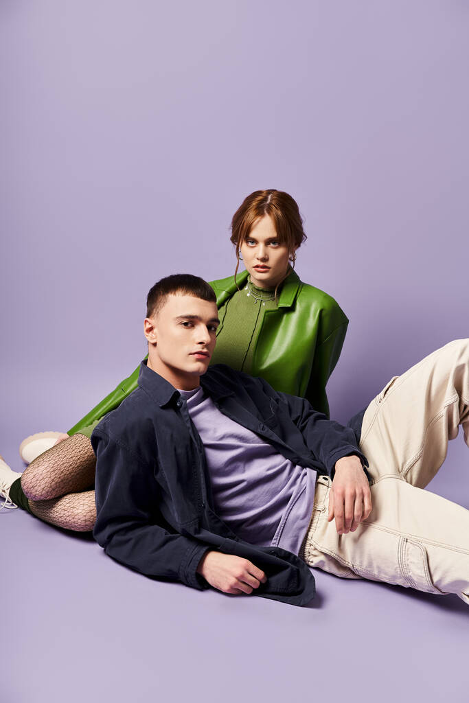 床に座って紫色の背景のカメラを見ている活気に満ちた服装のカップル - 写真・画像