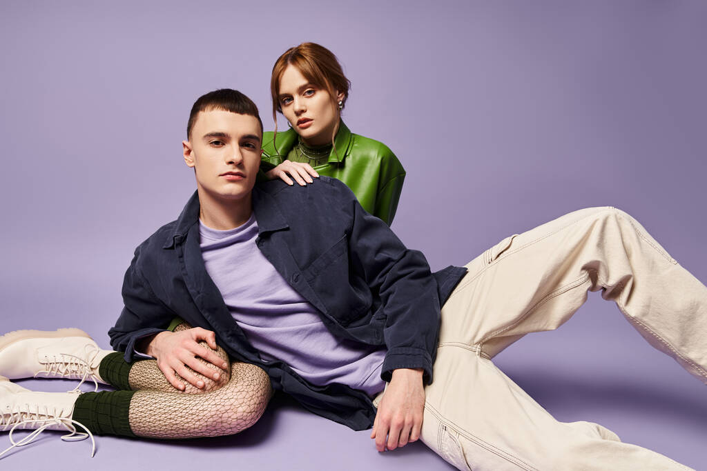 sophisticated couple in vibrant attires sitting on floor and looking at camera on purple backdrop - Photo, Image