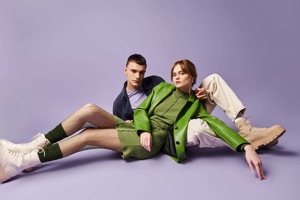 pareja de moda en trajes vibrantes sentado en el suelo y mirando a la cámara en el telón de fondo púrpura - Foto, Imagen