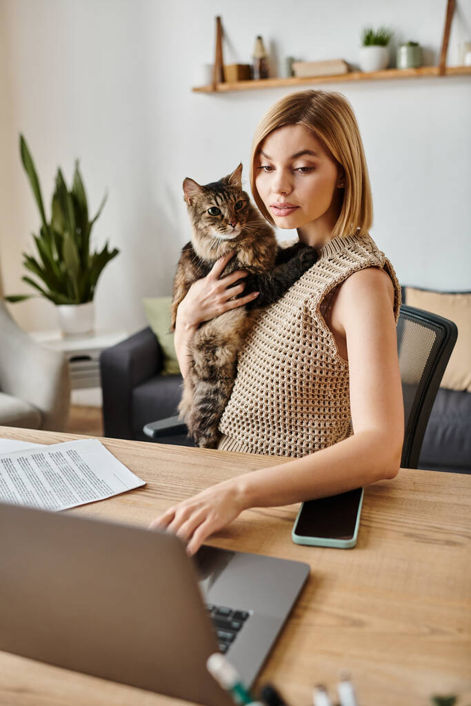 Stylowa kobieta z krótkimi włosami siedząca przy stole, pracująca na laptopie z kotem przy boku. - Zdjęcie, obraz