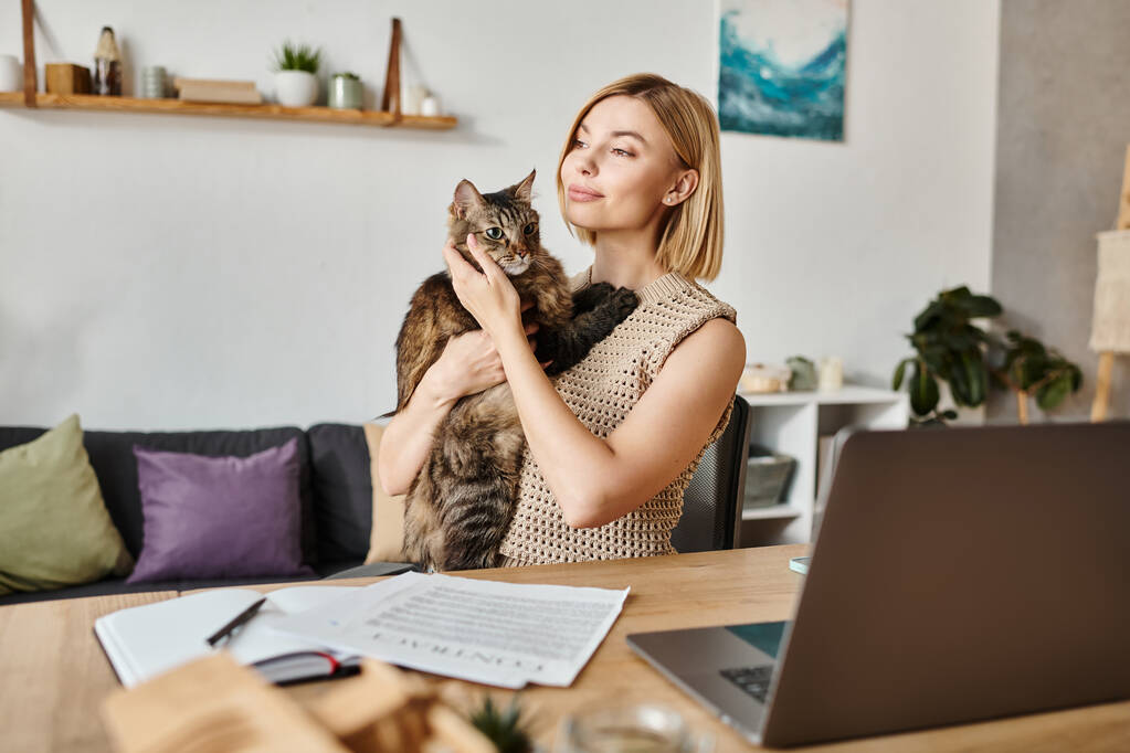Ніжна жінка з коротким волоссям, сидячи за столом, обережно тримає кішку в руках вдома. - Фото, зображення