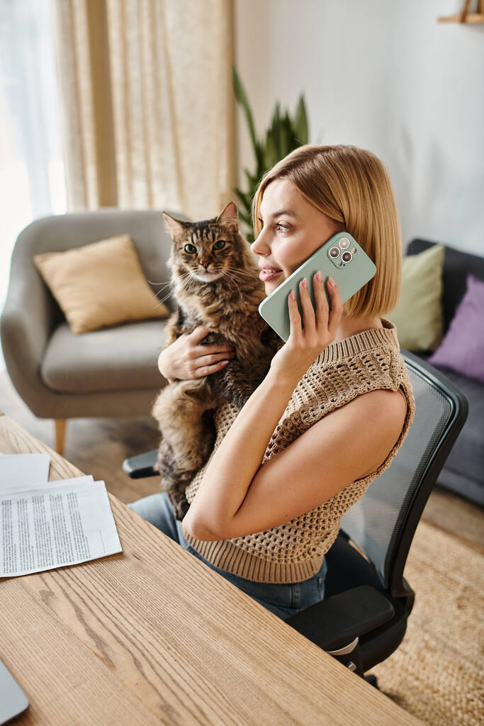 Una donna elegante con i capelli corti si siede a una scrivania, parlando al telefono con un simpatico gatto sdraiato sulle ginocchia. - Foto, immagini
