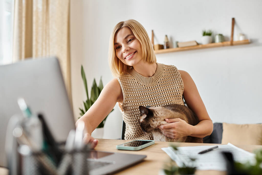 Una donna con i capelli corti si siede alla scrivania, assorta nel suo computer portatile mentre il suo gatto si siede accanto a lei, creando un'atmosfera tranquilla.. - Foto, immagini