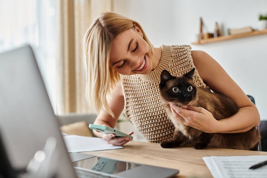 Una mujer con el pelo corto sentado en un escritorio, con amor sosteniendo un gato en sus brazos. - Foto, Imagen