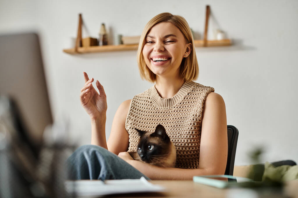 Una mujer con el pelo corto se sienta en un escritorio, interactuando pacíficamente con su gato en casa. - Foto, imagen
