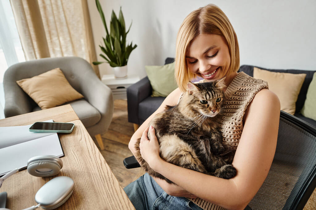 Lyhyttukkainen nainen pitää kissaansa rauhallisesti, kun hän istuu tuolilla kotona, muodostaen rauhallisen siteen.. - Valokuva, kuva