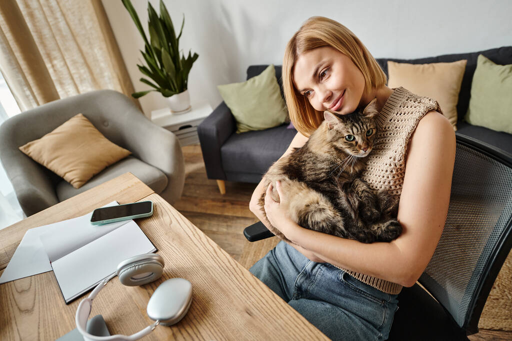 Kobieta z krótkimi włosami siedząca przy stole, trzymająca i głaskająca kota w spokojnej chwili w domu. - Zdjęcie, obraz