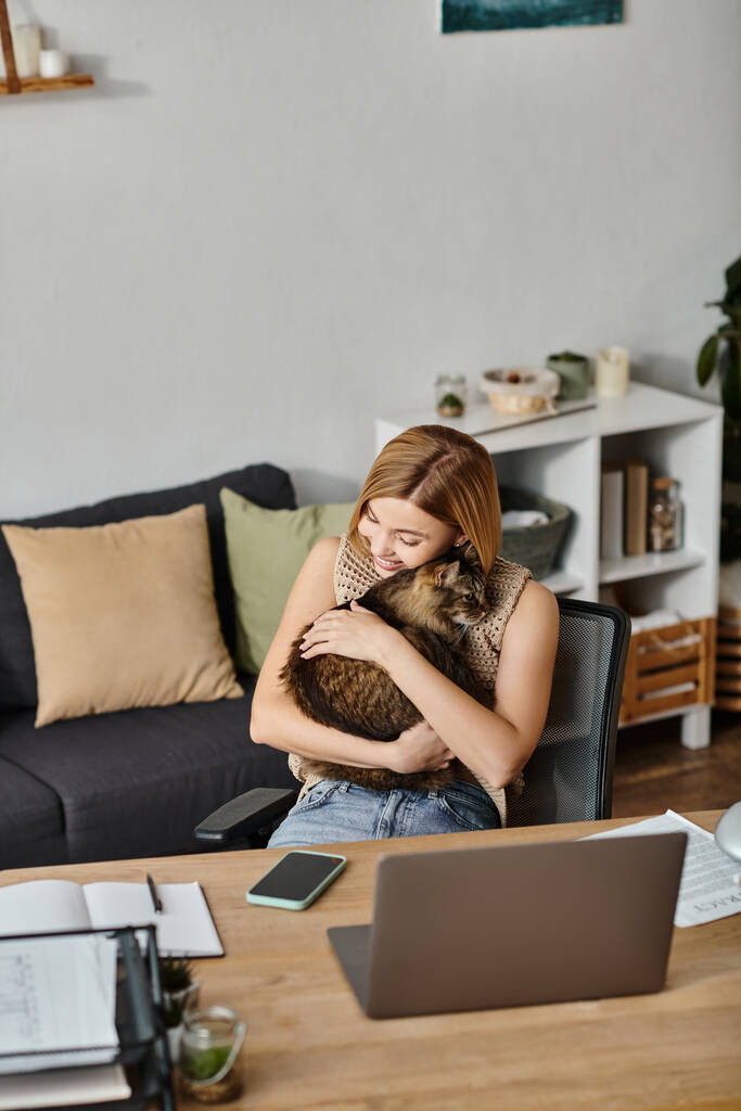 Egy rövid hajú, elegáns nő ül az íróasztalnál, szeretettel fogja a macskáját, és élvezi a pihenés pillanatát otthon.. - Fotó, kép