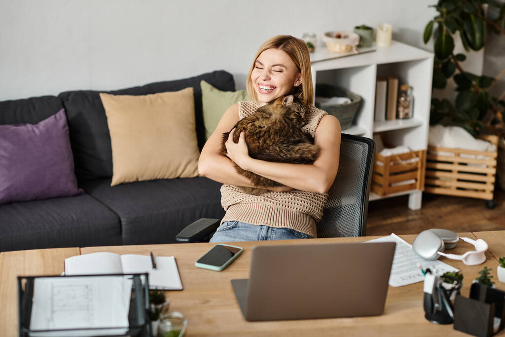 Κοντά μαλλιά γυναίκα κάθεται στο γραφείο, τρυφερά αγκαλιάζει μια χνουδωτή γάτα. - Φωτογραφία, εικόνα