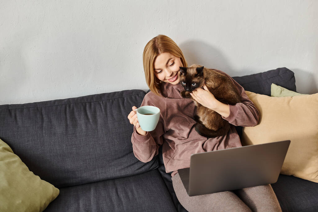 Una donna con i capelli corti si rilassa su un divano, tenendo un gatto tra le braccia mentre si gode una tazza di caffè. - Foto, immagini