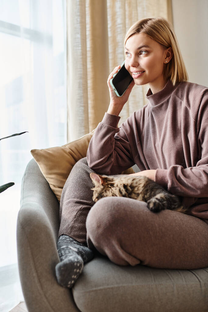 Uma mulher elegante com cabelo curto conversando em seu telefone celular, compartilhando um momento com seu gato adorável no sofá. - Foto, Imagem