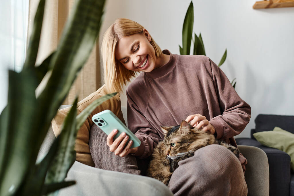 Kısa saçlı bir kadın kanepede oturuyor, bir kediyi nazikçe kollarına alıyor, şefkat gösteriyor ve birlikte huzurlu bir anın tadını çıkarıyor.. - Fotoğraf, Görsel