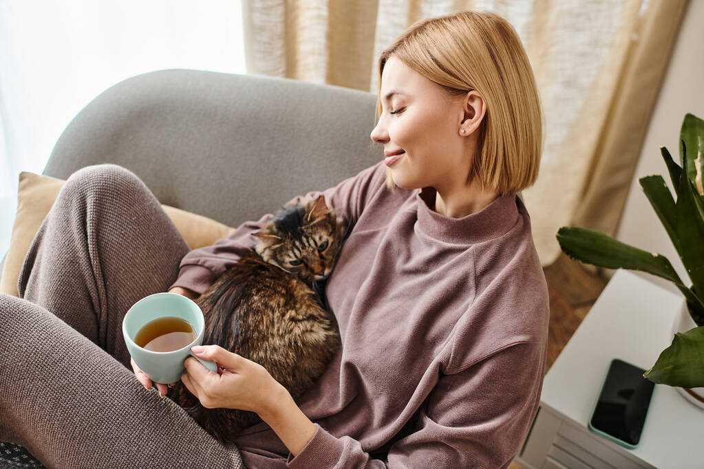 Μια κομψή γυναίκα σαλόνια σε έναν καναπέ, απολαμβάνοντας το τσάι και αγκαλιάζει μια γάτα περιεχόμενο σε μια γαλήνια εγχώρια σκηνή. - Φωτογραφία, εικόνα