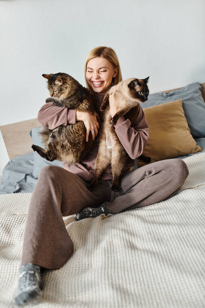 A serene woman with short hair sits on a bed, holding two cats close to her, enjoying a peaceful moment at home. - Photo, Image
