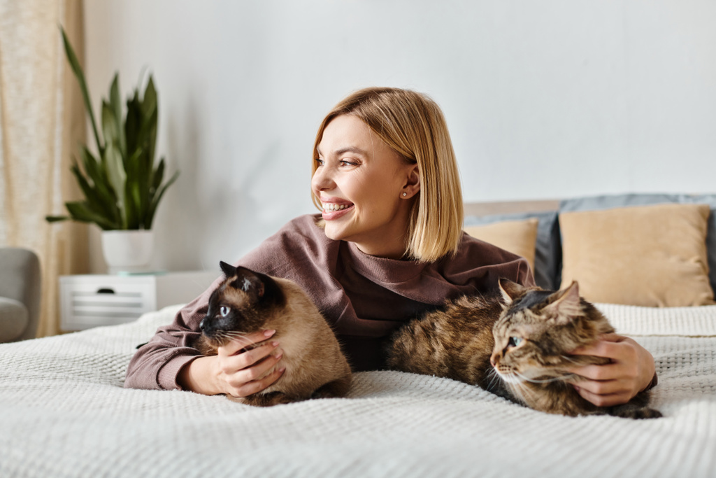 Μια γυναίκα με κοντά μαλλιά χαλαρώνει σε ένα κρεβάτι με δύο γάτες στο πλευρό της, απολαμβάνοντας μια ειρηνική στιγμή στο σπίτι. - Φωτογραφία, εικόνα