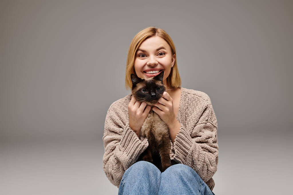 Okouzlující žena s krátkými vlasy sedí na podlaze, drží svou milovanou kočku, sdílí klidný moment doma. - Fotografie, Obrázek
