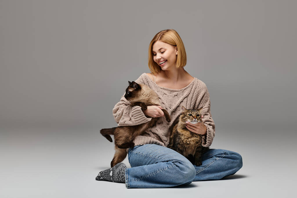 Μια γυναίκα με κοντά μαλλιά κάθεται στο πάτωμα, με αγάπη κρατώντας δύο γάτες σε μια γαλήνια και ειρηνική στιγμή στο σπίτι. - Φωτογραφία, εικόνα