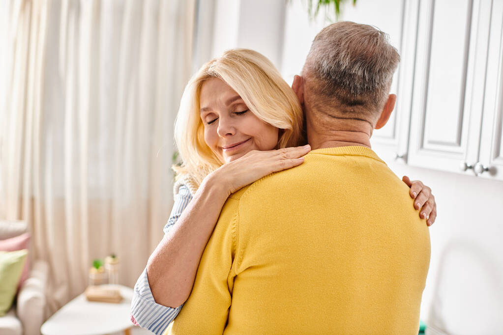 Een volwassen vrouw in gezellige huiskleding knuffelt een man in een warme, liefdevolle omhelzing in hun woonkamer. - Foto, afbeelding