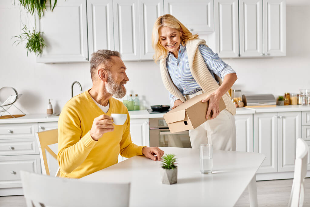 Um homem e uma mulher maduros em roupa de casa aconchegante são vistos movendo caixas em uma cozinha juntos em casa. - Foto, Imagem