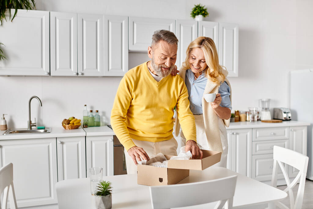 Ένας ώριμος άντρας και μια γυναίκα με άνετα ρούχα ανοίγουν ένα κουτί σε μια ζεστή κουζίνα, μοιράζονται μια στιγμή περιέργειας και προσμονής.. - Φωτογραφία, εικόνα