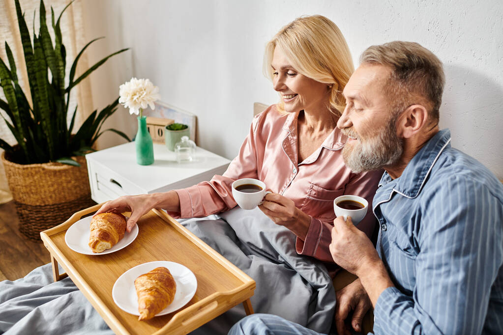 Una pareja madura en acogedora ropa de casa sentada en un sofá, bebiendo café y disfrutando de pasteles juntos en un ambiente cálido y acogedor. - Foto, imagen