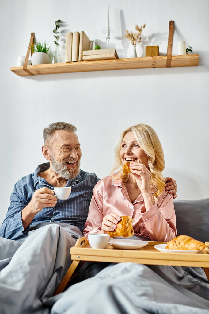 Un hombre y una mujer maduros disfrutan de una comida mientras están sentados en un cómodo sofá en su acogedor dormitorio, vestidos con ropa de casa. - Foto, Imagen