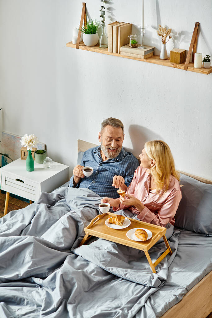 Ein reifer Mann und eine reife Frau in gemütlicher Hausmannskleidung sitzen zusammen auf einem Bett und teilen einen ruhigen Moment der Zweisamkeit. - Foto, Bild
