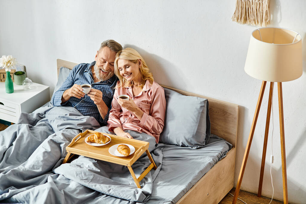 Зріла любляча пара в затишному домашньому одязі сидить на ліжку, насолоджуючись один одним - Фото, зображення