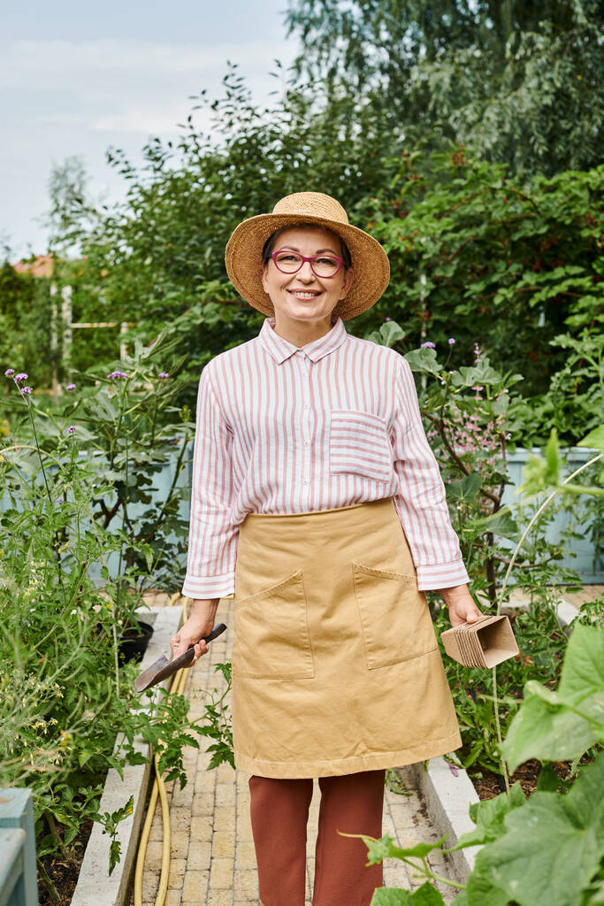 hermosa mujer madura alegre en sombrero y atuendo casual sosteniendo herramientas de jardinería y sonriendo a la cámara - Foto, imagen