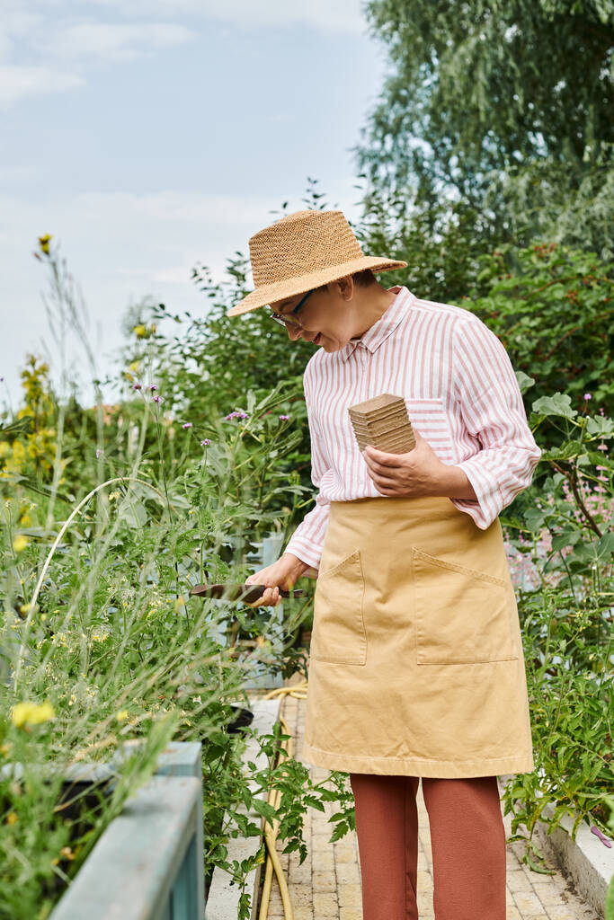 mulher madura alegre atraente com chapéu de palha usando suas ferramentas de jardinagem e sorrindo alegremente - Foto, Imagem