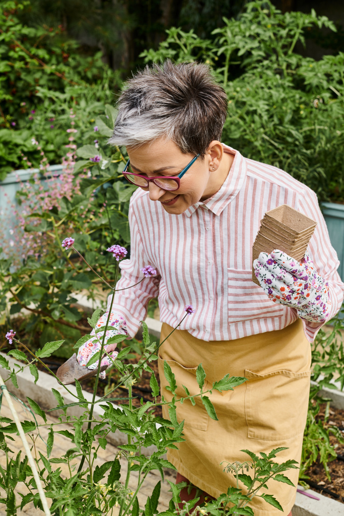 mulher madura alegre atraente com óculos e luvas usando equipamentos de jardinagem em suas flores - Foto, Imagem