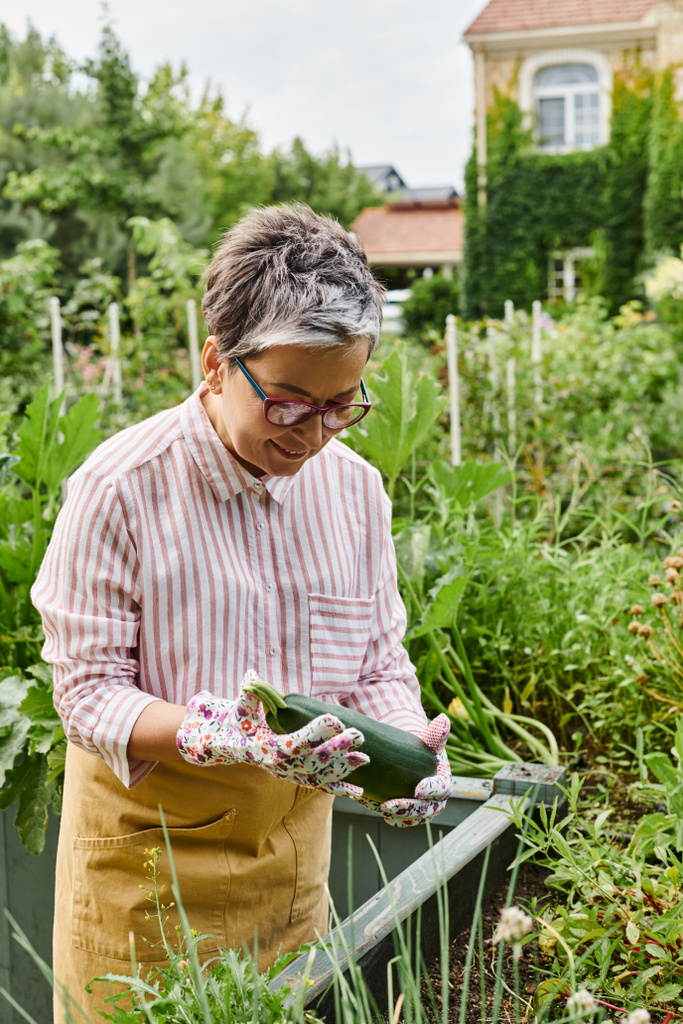 приваблива весела зріла жінка в повсякденному вбранні з окулярами, що тримає свіжі кабачки в своєму саду - Фото, зображення