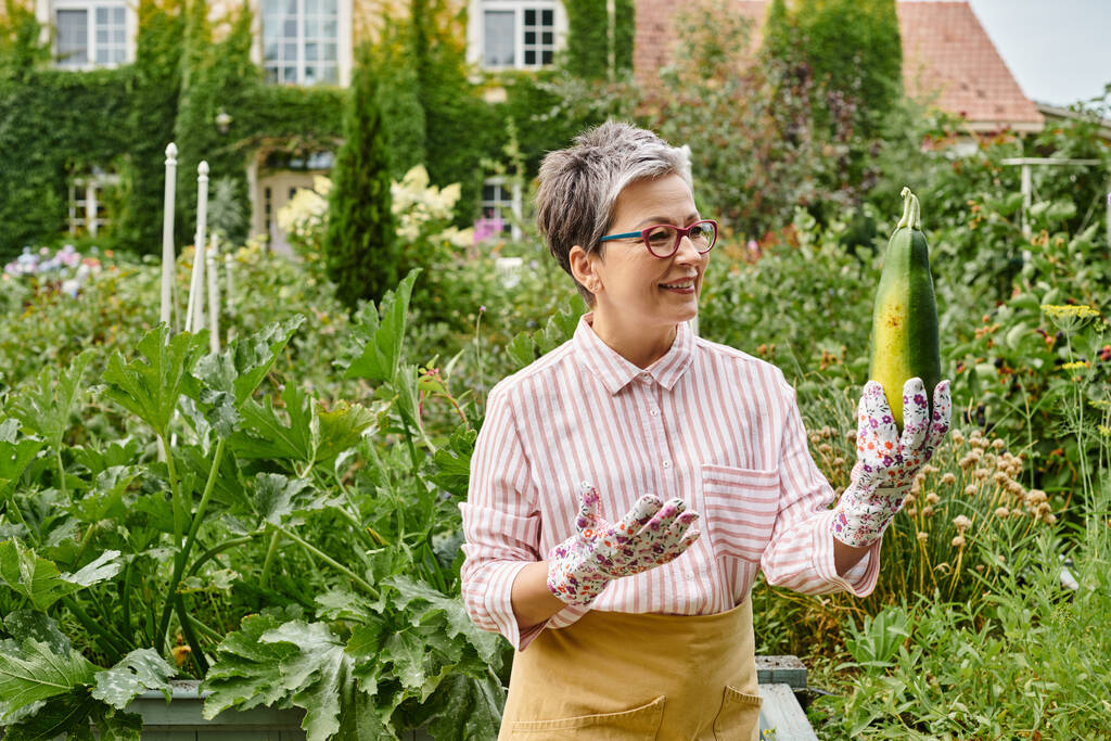 attraktive fröhliche reife Frau in lässiger Kleidung mit Gläsern, die frische Zucchini in ihrem Garten halten - Foto, Bild