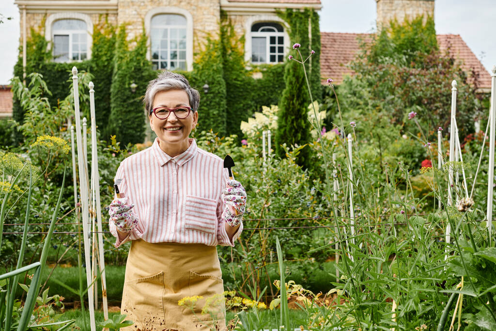 cheerful mature woman with gloves and glasses holding gardening tools in hands and smiling at camera - Photo, Image