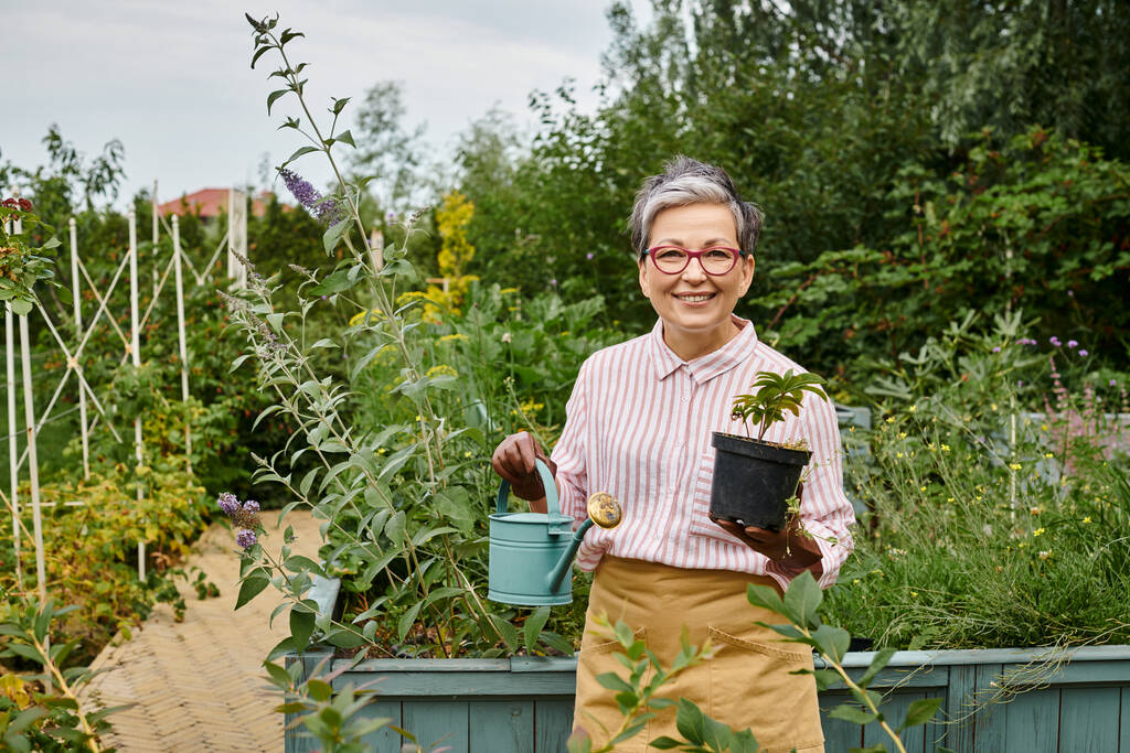 atractiva mujer madura alegre sosteniendo regadera y olla con planta y sonriendo a la cámara, Inglaterra - Foto, imagen