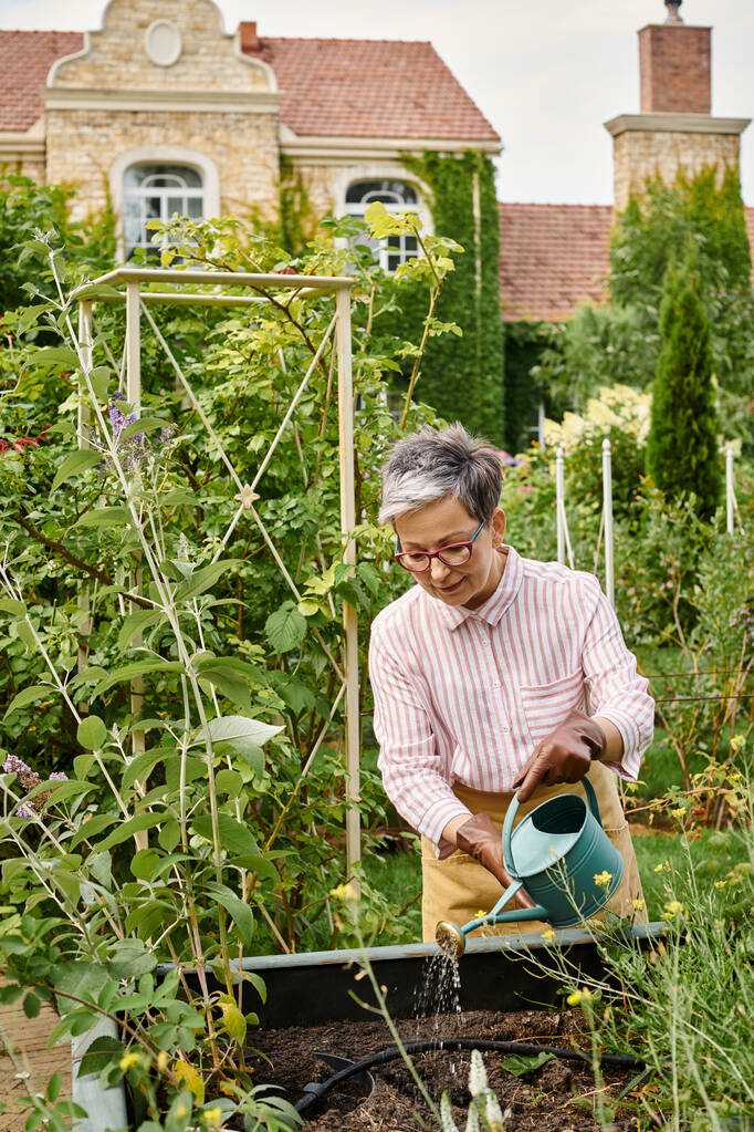 привлекательная веселая зрелая женщина в стильных очках поливает растения в своем саду в Англии - Фото, изображение