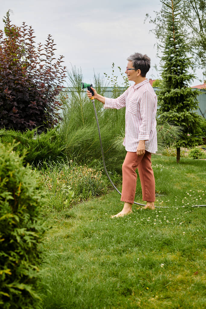 гарний вигляд весела зріла жінка в окулярах, використовуючи шланг, щоб поливати її живі рослини в її саду - Фото, зображення