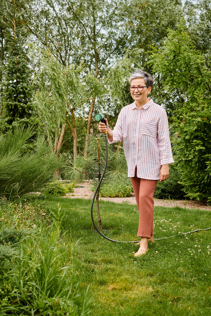 bella cercando allegra donna matura con gli occhiali utilizzando tubo per innaffiare le sue piante vivaci nel suo giardino - Foto, immagini