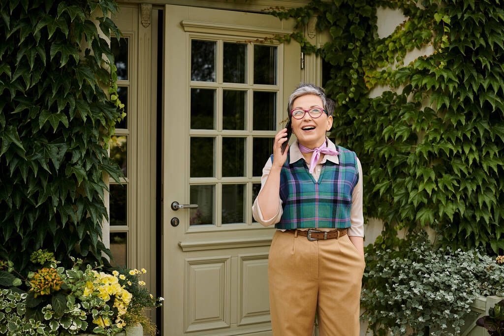 ώριμη χαρούμενη κομψή γυναίκα με γυαλιά και κοντά μαλλιά μιλώντας από το τηλέφωνό της κοντά στο σπίτι στην Αγγλία - Φωτογραφία, εικόνα